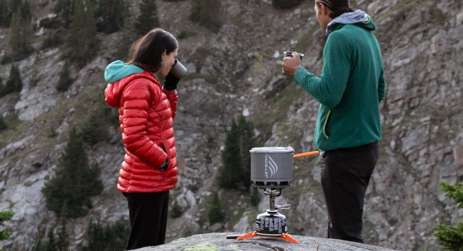 a couple in red and green jackets drink from camp mugs while a jetboil stove sits on a rock in the foreground. 